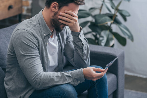 грустный мужчина сидит на диване, держит фото и плачет дома, скорбящее расстройство концепции
