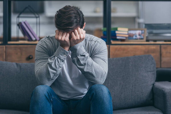 man sitting on couch, crying and and covering face with hands at home