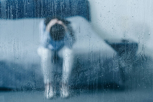 подавленная женщина сидит на кровати и держит голову в руках через окно с каплей дождя
