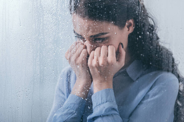 depressed woman covering face with hands and crying at home through window with raindrops