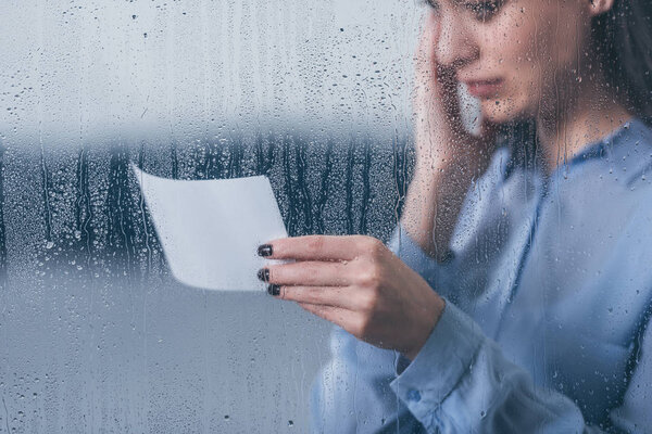 cropped view of grieving woman holding photograph and crying through window with raindrops