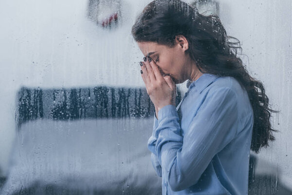 depressed woman covering face with hands and crying at home through window with raindrops and copy space