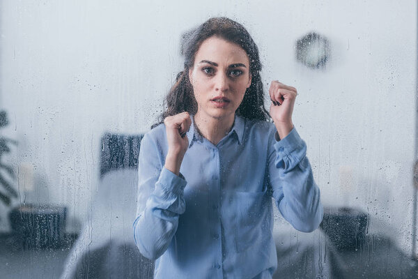 beautiful sad woman with clenched fists looking at camera through window with raindrops