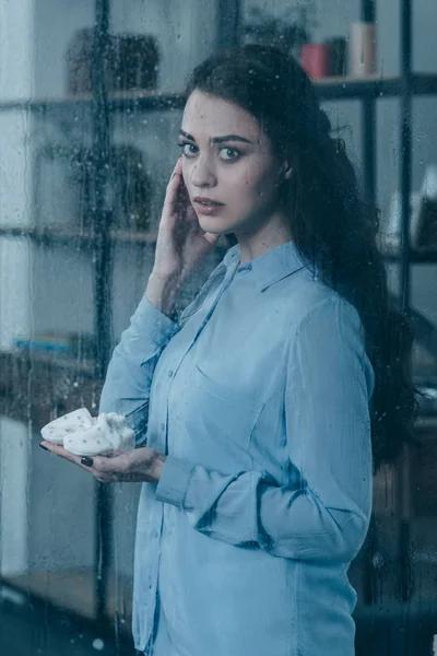 ベビー シューズを押しながら雨で窓からカメラを見て悲嘆の母 — ストック写真