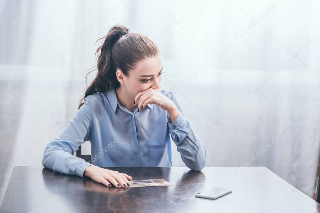 upset woman in blue blouse sitting at table with photo and thinking in room, grieving disorder concept