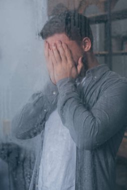 yüzünü elleriyle kapsayan ve yağmur damlaları ile pencereden ağlıyor yetişkin adam