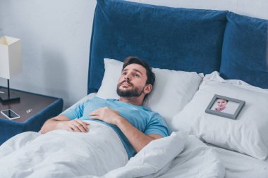 depressed man lying in bed near photo frame with picture of woman on pillow at home clipart