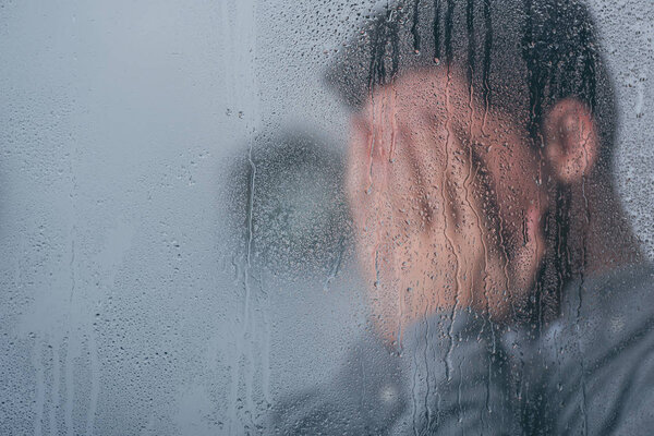 selective focus of raindrops on window with man covering face and crying on background