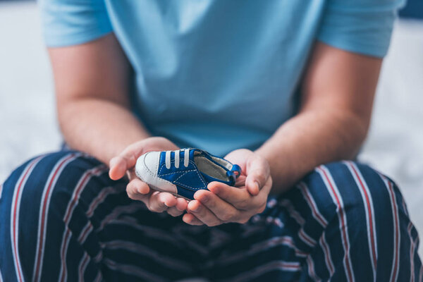 cropped view of man sitting on bed and holding baby shoe