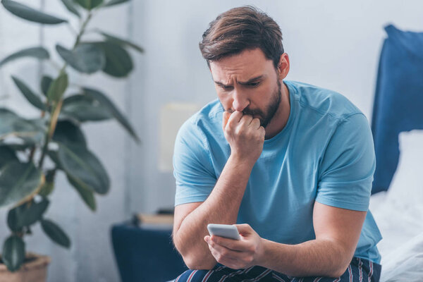 upset man sitting on bed, covering mouth with hand and using smartphone at home