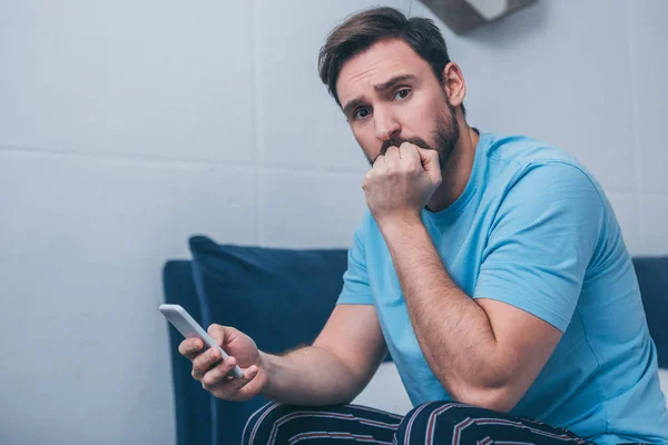 upset man sitting on bed, covering mouth with hand and using smartphone at home