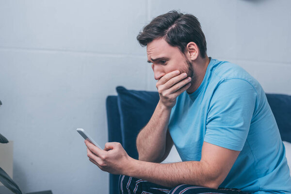 grieving man sitting on bed, covering mouth with hand and using smartphone at home