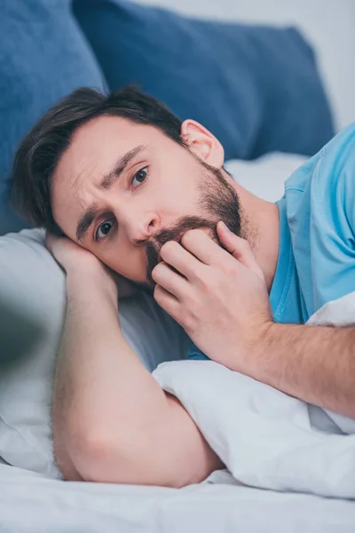 scared man lying in bed, looking at camera and covering mouth with hand
