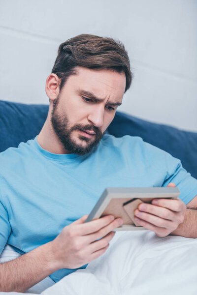 selective focus of sad handsome man lying in bed, looking at photo frame and grieving at home