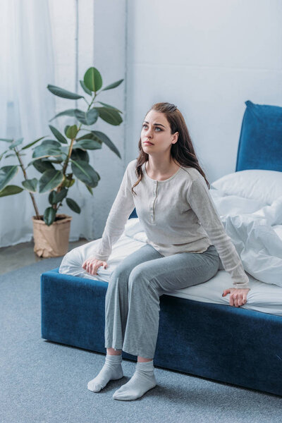 sad woman in pajamas looking up while sitting on bed at home