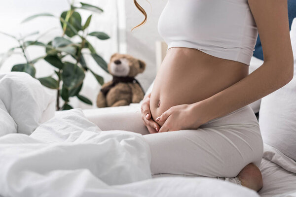 Partial view of pregnant woman touching belly while sitting on bed