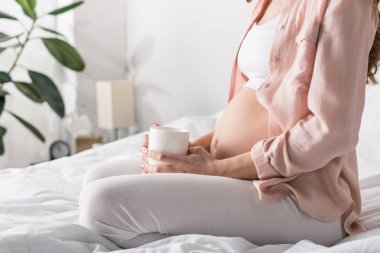 Hamile kadın kahve fincanı ile yatakta oturuyordu görünümünü kırpılmış
