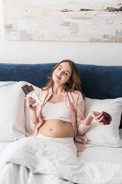 チョコレート バーと目を閉じてアップルが保持している夢のような妊娠中の女性 — ストック写真