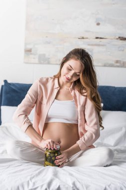 Pregnant woman sitting on bed and opening jar with pickles clipart