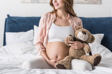 Yatağın üstüne oturan ve oyuncak ayı tutan hamile kadın kısmi görünümü