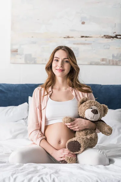 熊のぬいぐるみでベッドの上に座って陽気な妊娠中の女性 — ストック写真