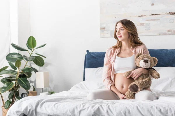 ベッドの上に座っているクマのぬいぐるみを持つ夢のような妊娠中の女性 — ストック写真