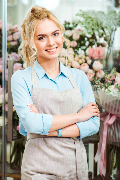 beautiful smiling female florist in apron looking at camera with flower shop on background