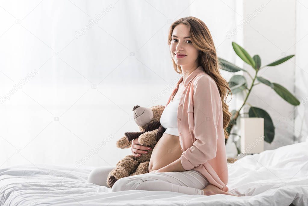Charming pregnant woman sitting on bed with teddy bear