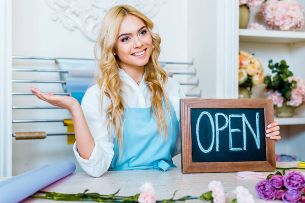 beautiful smiling female flower shop owner gesturing with hand and holding chalkboard with 'open' lettering 