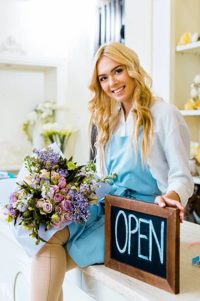 beautiful smiling female flower shop owner holding chalkboard with \'open\' lettering and colorful bouquet while looking at camera