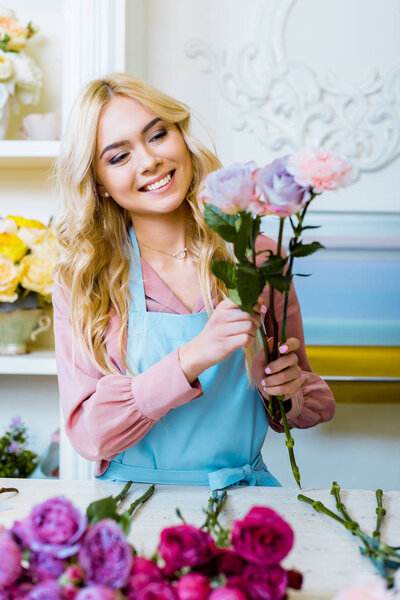 beautiful happy female florist in apron arranging bouquet with roses in flower shop 