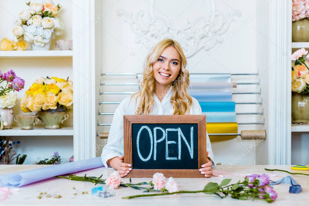 beautiful smiling female flower shop owner looking at camera and holding chalkboard with 'open' lettering 