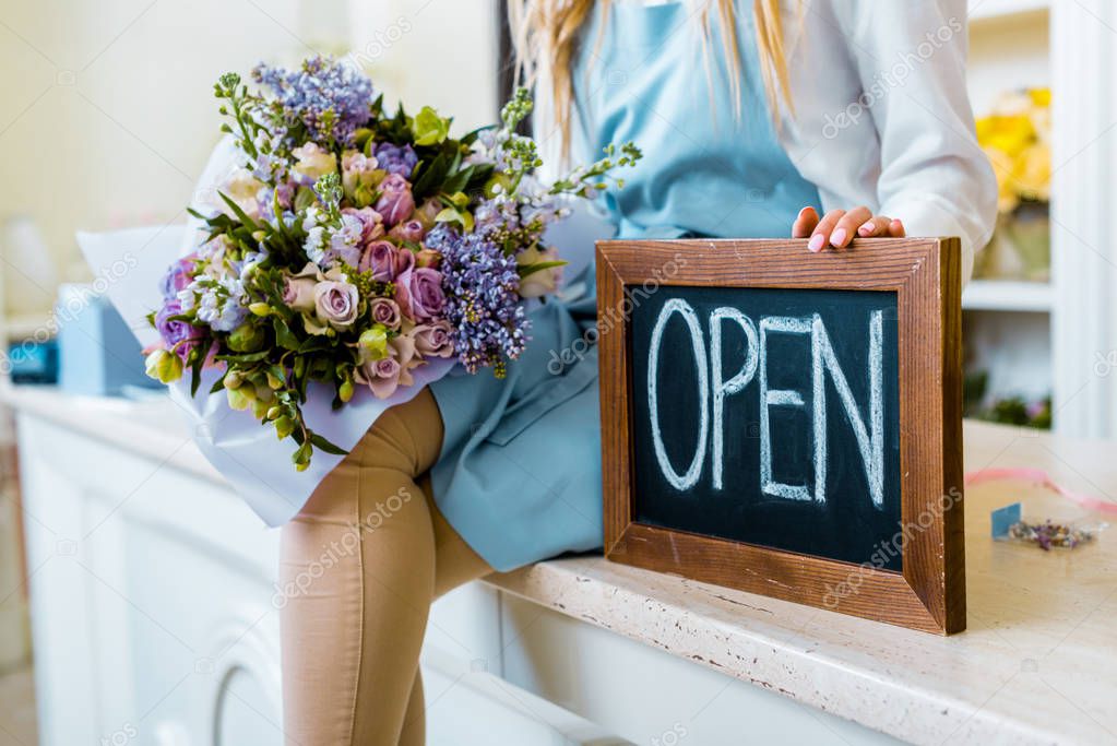 cropped view of female flower shop owner holding chalkboard with 'open' lettering and colorful bouquet