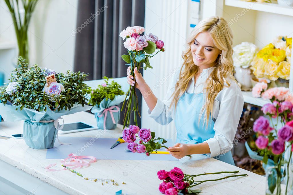 beautiful smiling female florist in apron arranging bouquet with roses in flower shop 