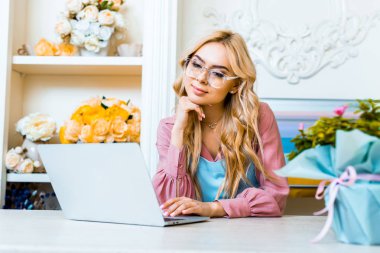 beautiful pensive female florist in glasses using laptop in flower shop clipart