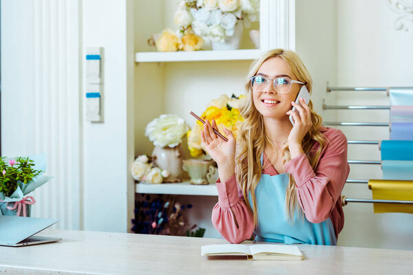 beautiful female flower shop owner in glasses talking on smartphone and holding pencil