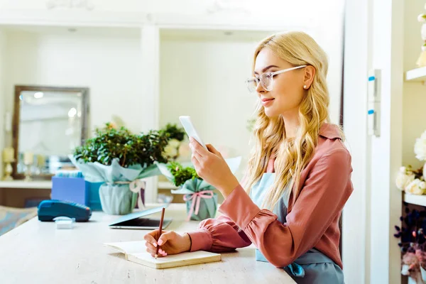 Beautiful Female Flower Shop Owner Glasses Writing Notebook Using Smartphone — Stock Photo, Image