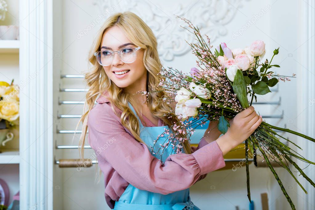 beautiful female florist in glasses holding bouquet in flower shop and looking away