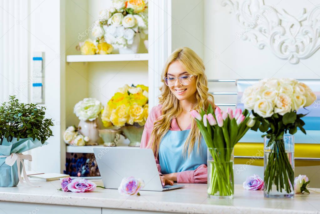 beautiful smiling female flower shop owner in glasses sitting at desk and using laptop