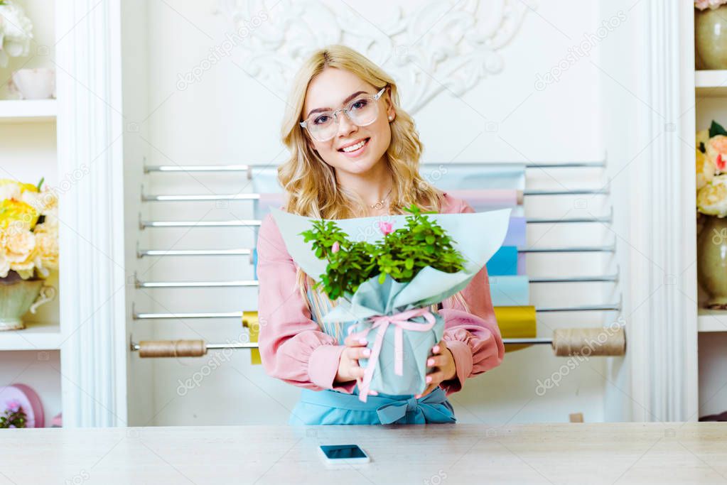 beautiful female florist in glasses looking at camera and presenting bouquet in flower shop