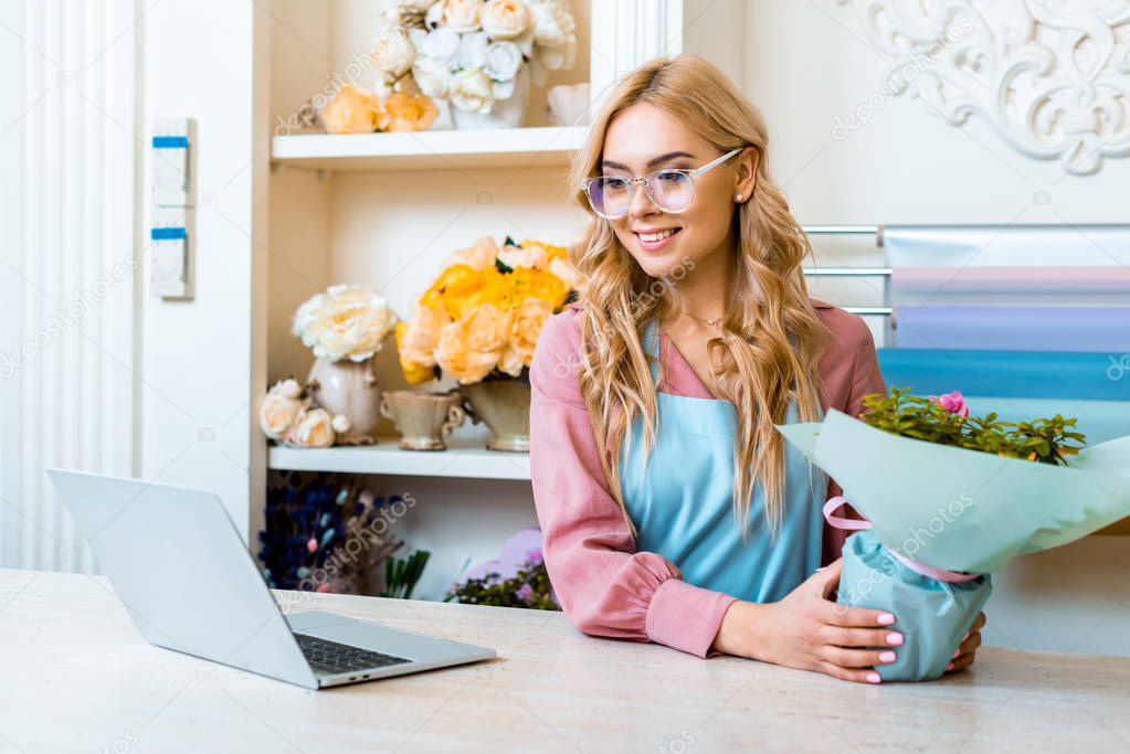 beautiful female florist in glasses sitting with floral bouquet at computer desk in flower shop