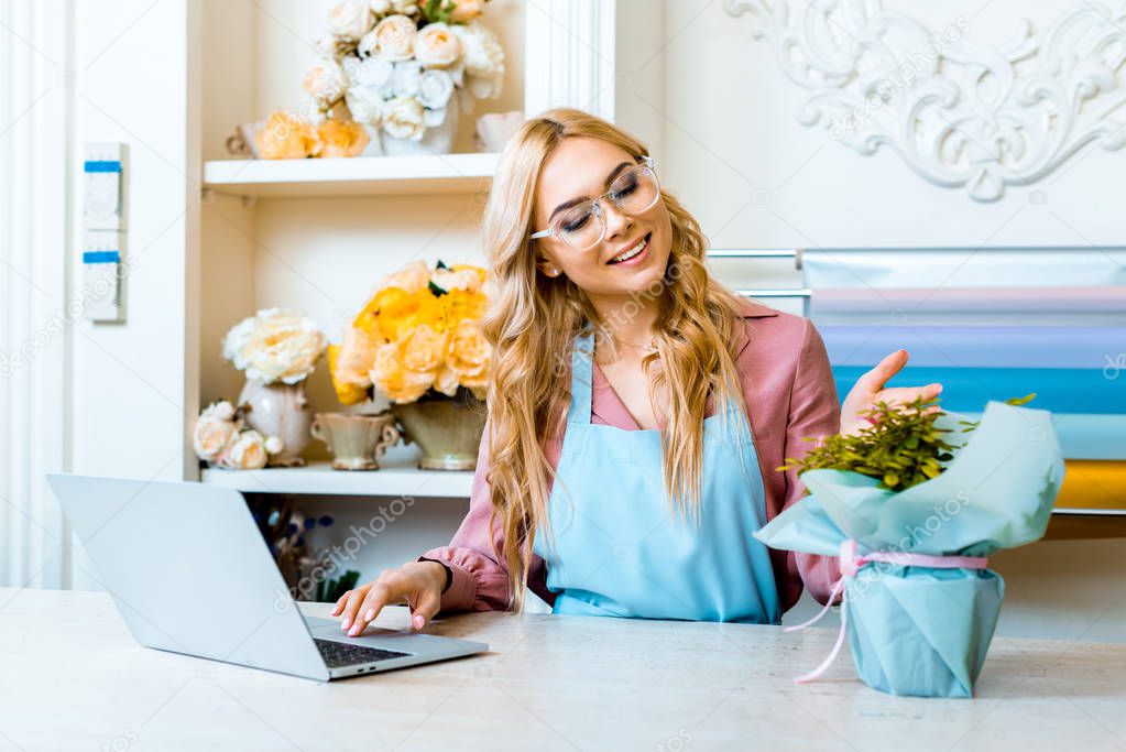 beautiful happy female florist in glasses sitting with floral bouquet at computer desk in flower shop