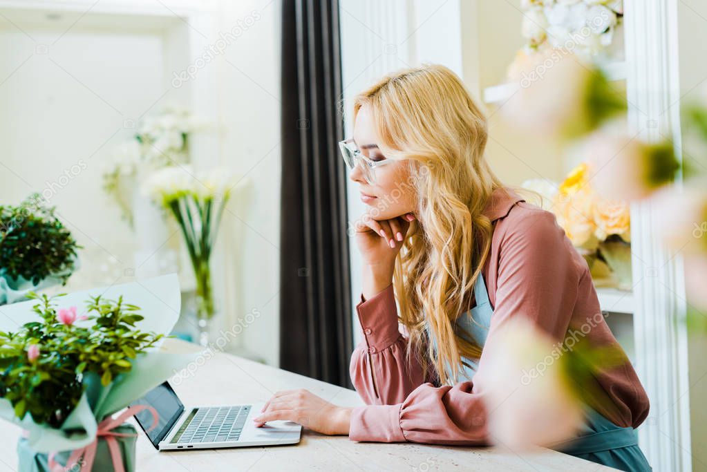 beautiful pensive female florist in glasses using laptop in flower shop