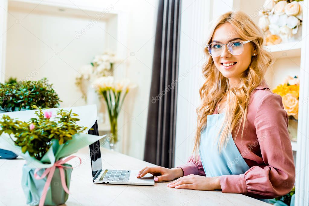 beautiful female florist in glasses using laptop in flower shop and looking at camera