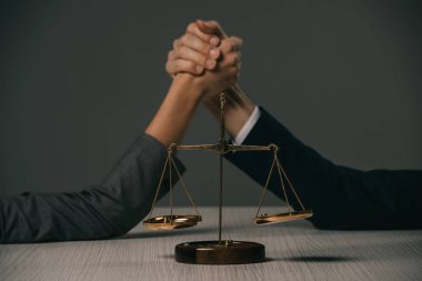 cropped view of businesspeople arm wretsling on wooden table with scales of justice on grey clipart