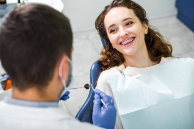 selective focus of cheerful woman in braces during examination of teeth near dentist clipart