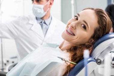selective focus of beautiful woman in braces during examination of teeth near dentist clipart