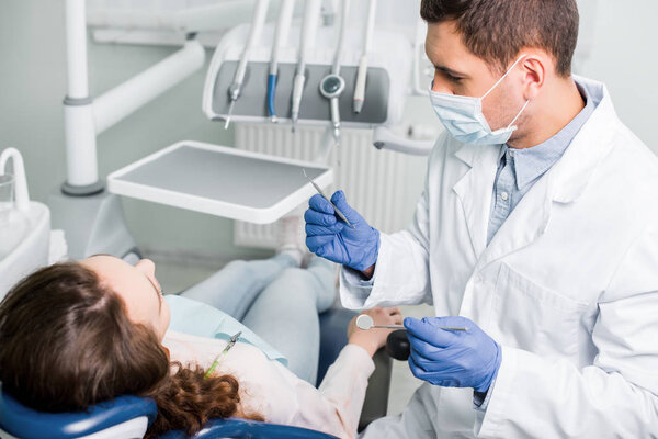 serious dentist in latex gloves and mask holding dental equipment near female patient 