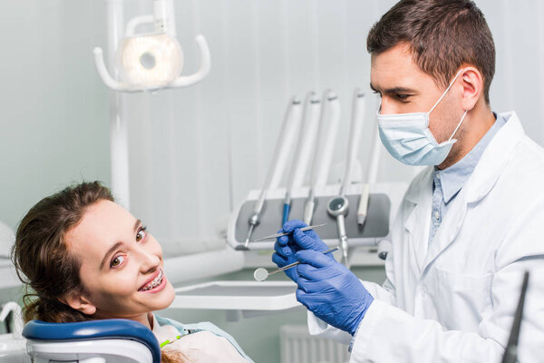 dentist in latex gloves and mask holding dental equipment near beautiful woman with braces 