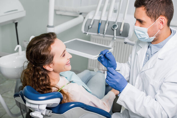 dentist in mask and latex gloves standing near smiling woman in dental clinic